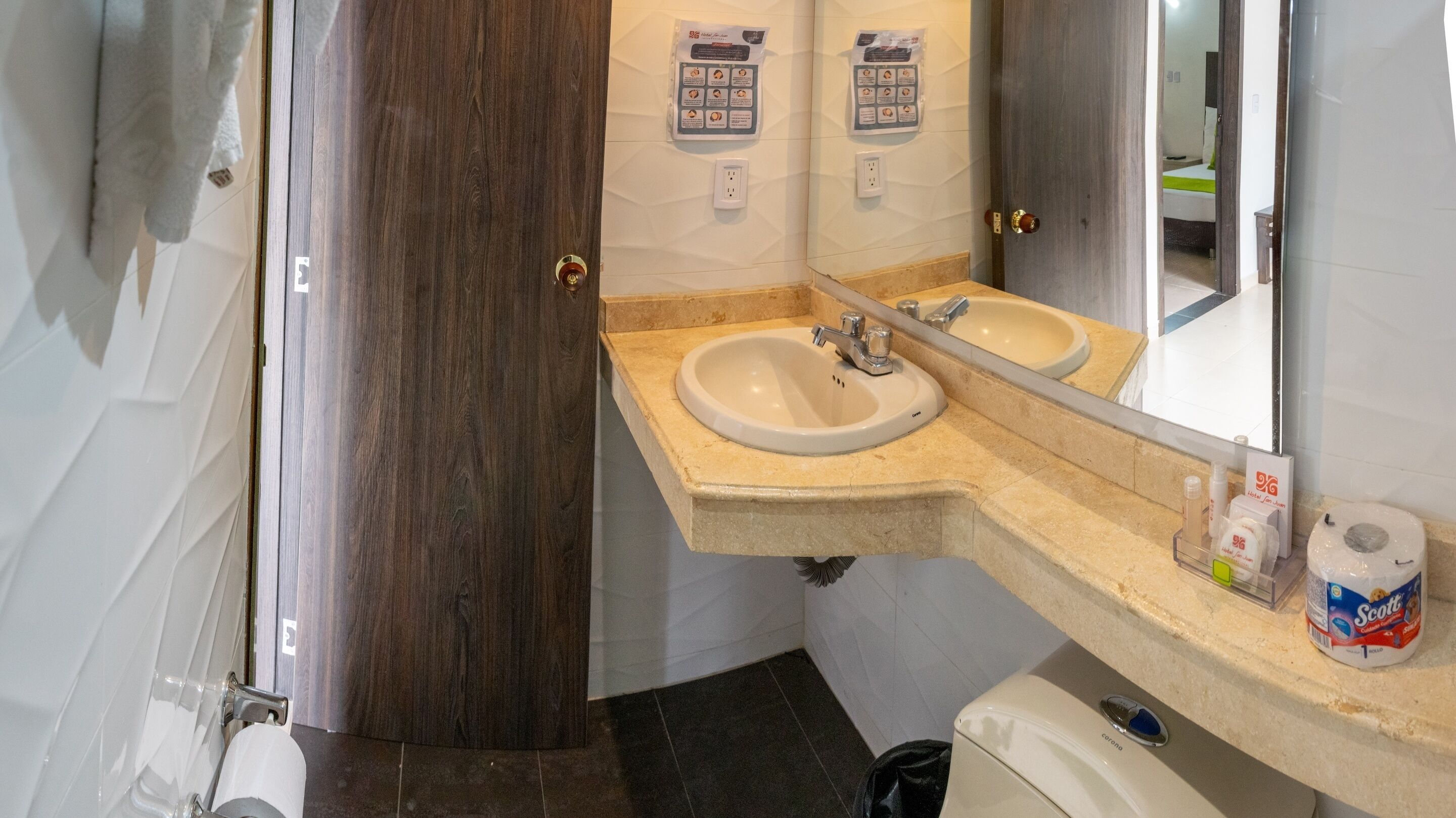 Une salle de bain avec deux lavabos, un miroir et des toilettes. On trouve des articles de toilette sur le comptoir et une serviette est accrochée au mur.