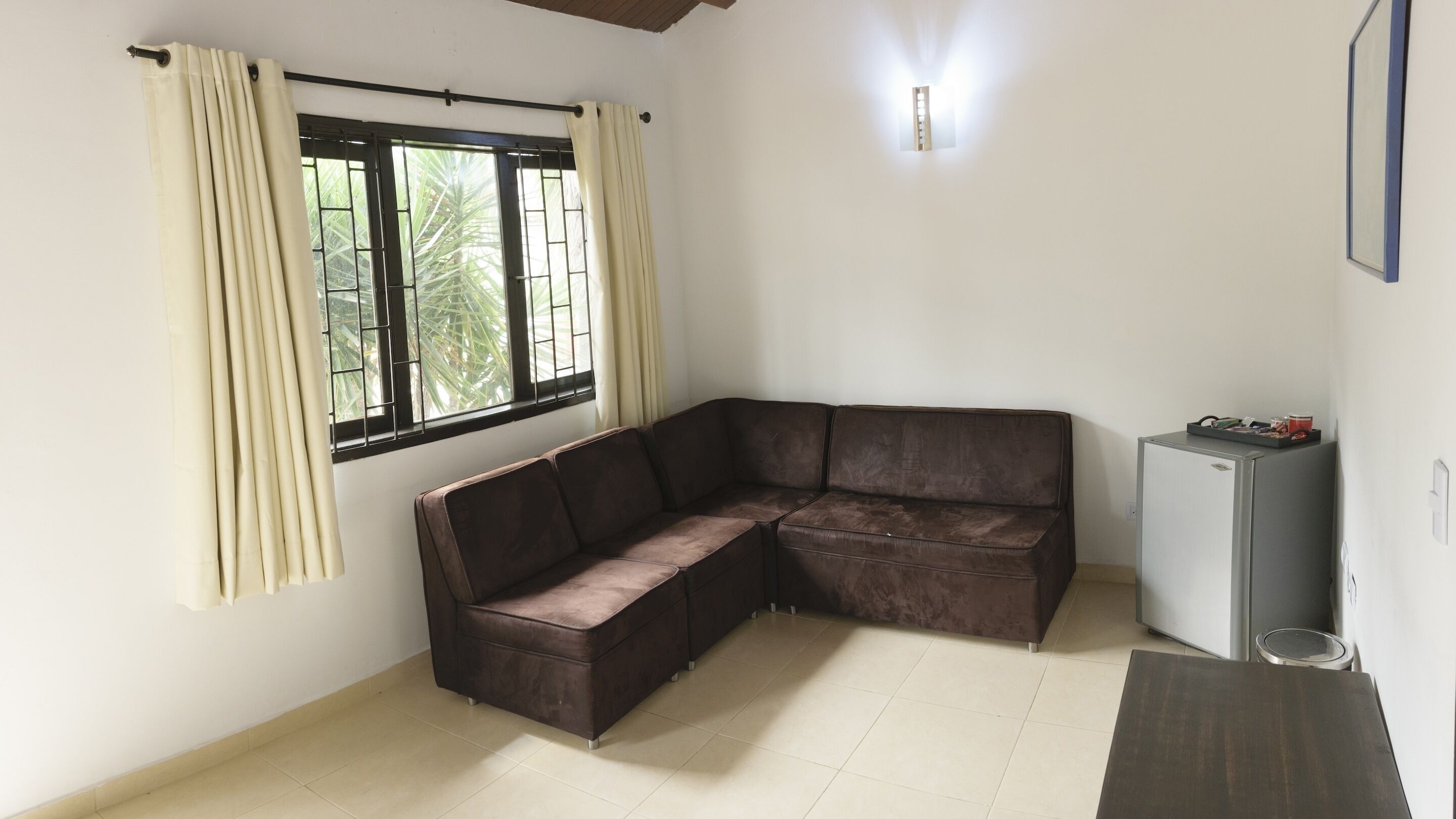 A room with a brown sectional sofa, a small refrigerator, and a window with curtains.