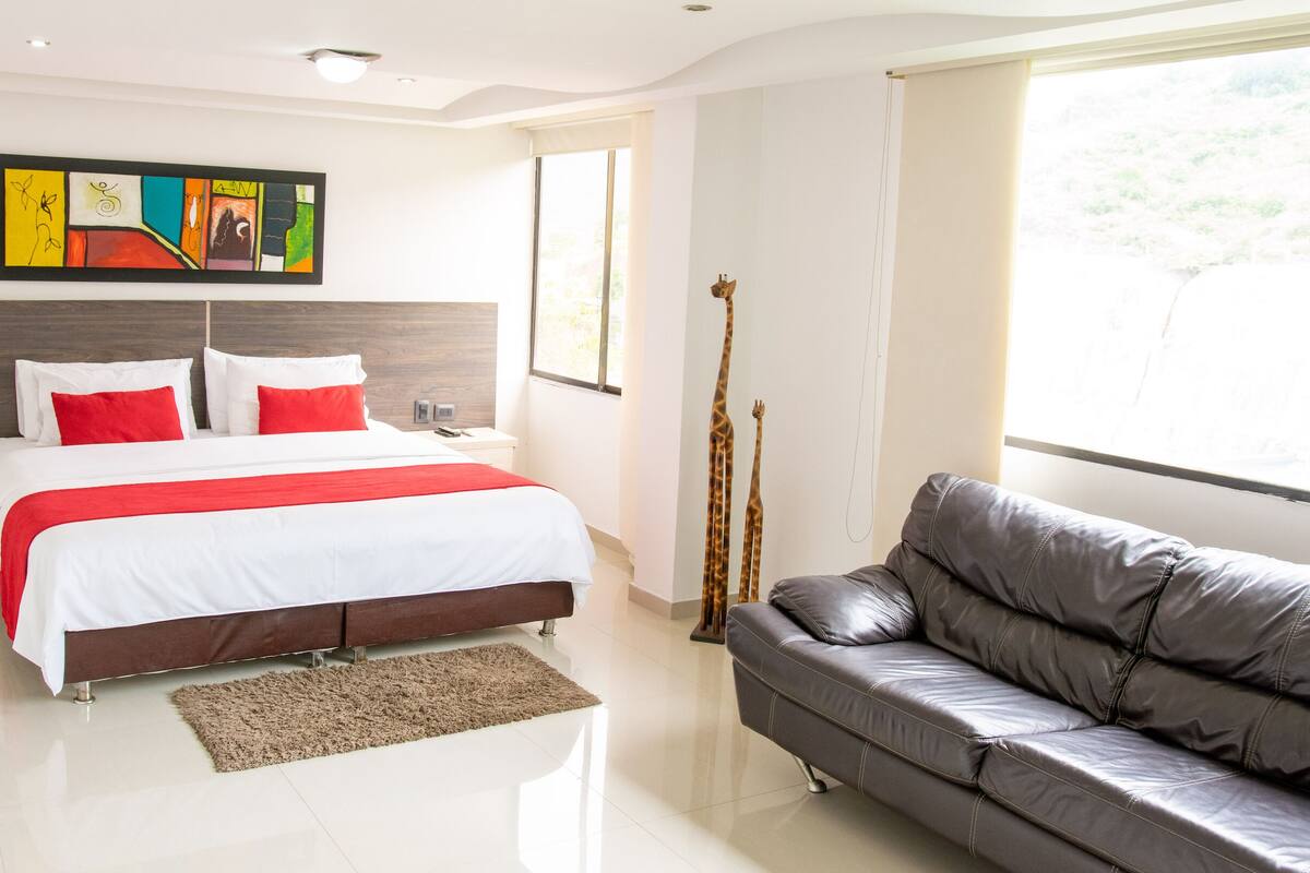 Een moderne slaapkamer met een groot bed, een grijze bank, een houten hoofdeinde, een kleurrijk abstract schilderij en een raam met uitzicht.