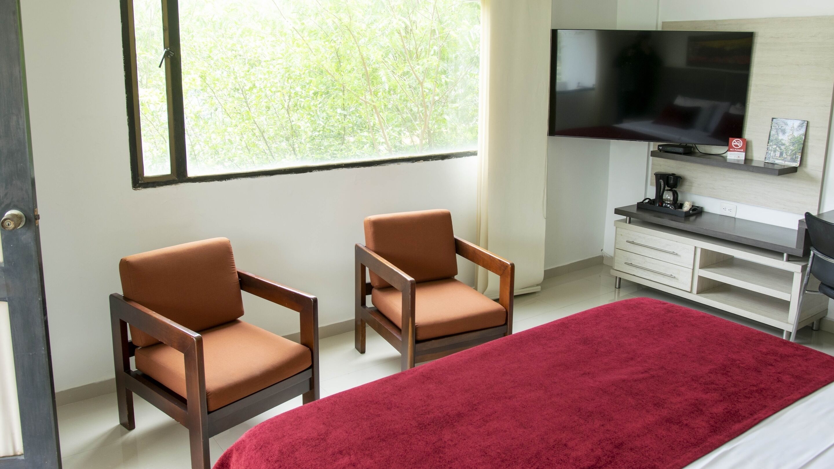 A hotel room with a bed, two armchairs, a TV, a desk, and a window with a view of trees.