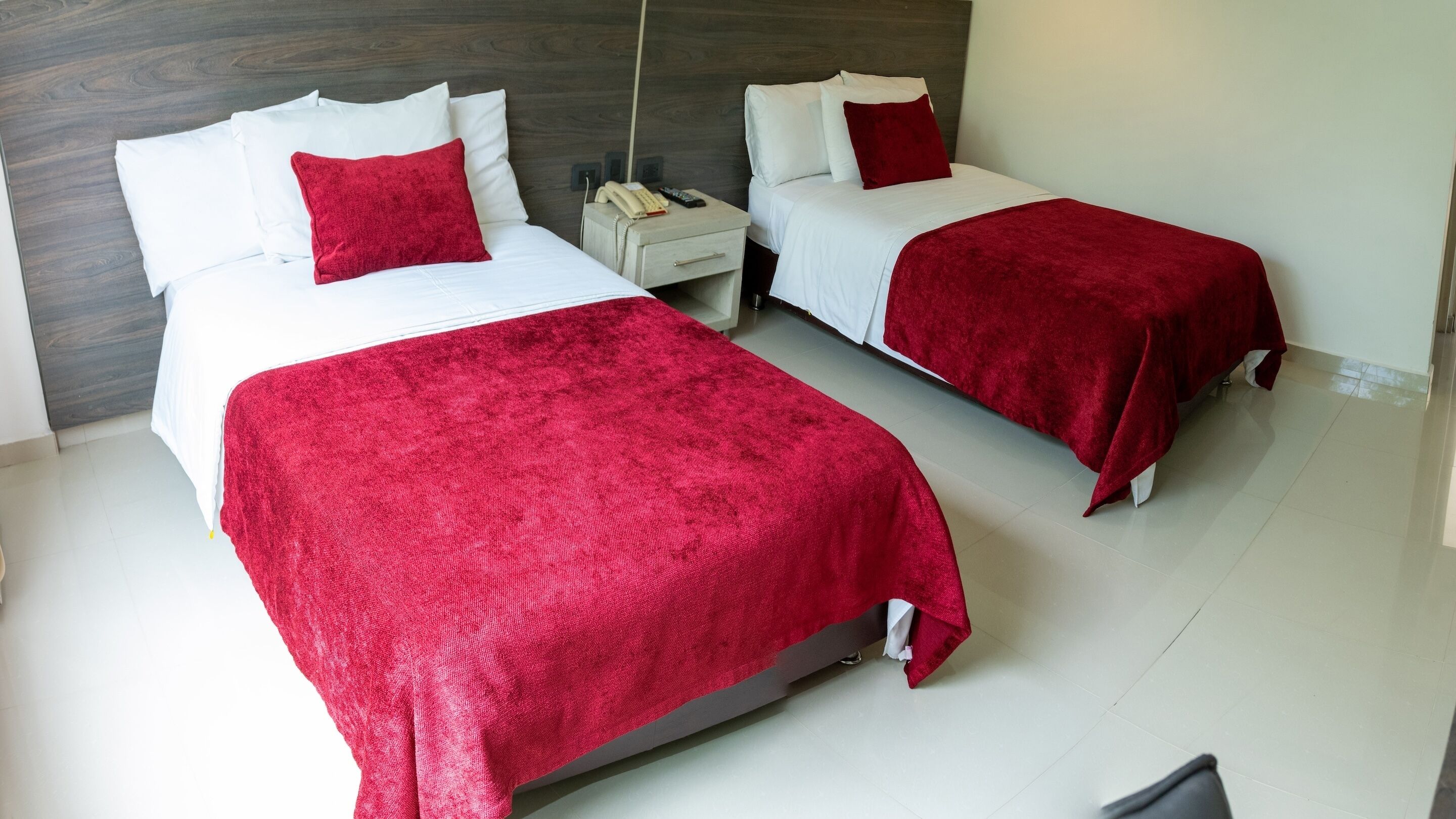 A hotel room with two beds, each with white bedding and red throw pillows. There is a nightstand with a telephone and remote controls.