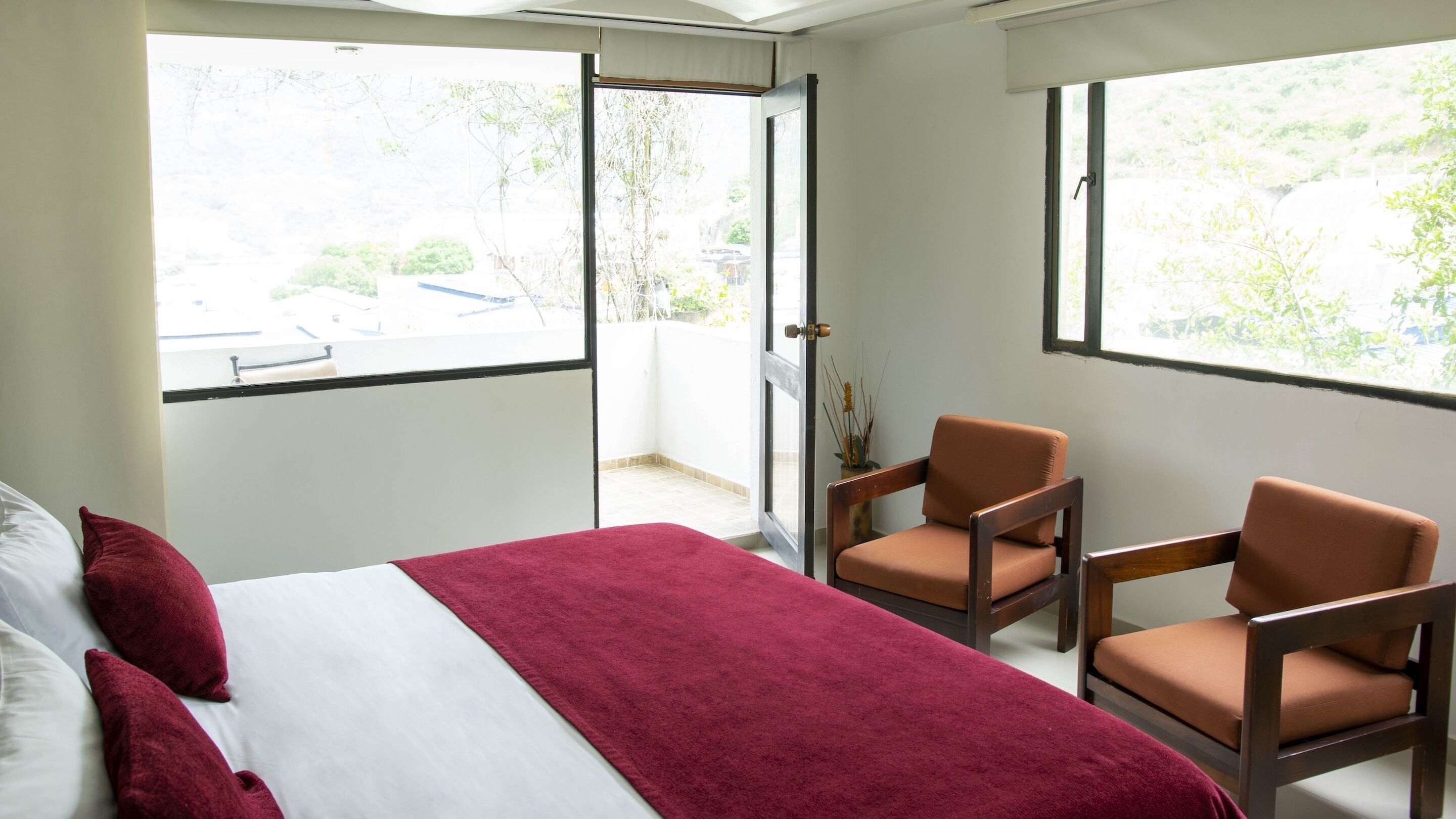 Habitación de hotel con cama, dos sillas, una manta roja y ventanas amplias que dan a zonas verdes y un edificio.