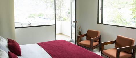 Habitación de hotel con cama, dos sillas, una manta roja y ventanas amplias que dan a zonas verdes y un edificio.