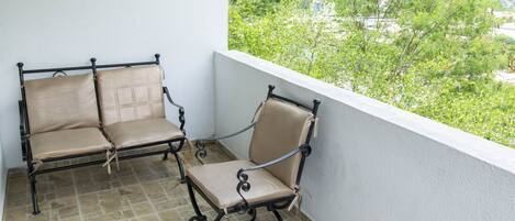 A balcony with two beige cushioned chairs and a small table, featuring a view of greenery and a bridge in the background.
