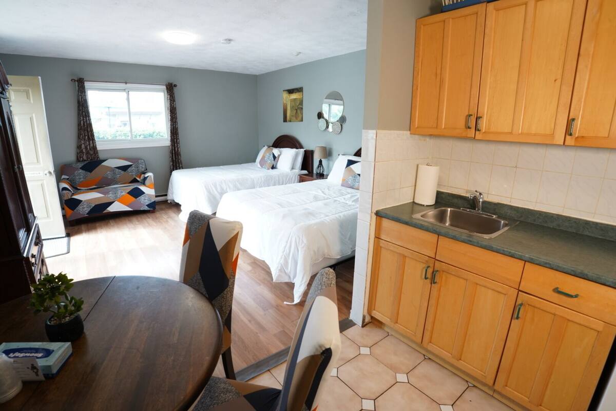 A hotel room with two beds, a dining table, a kitchen area with wooden cabinets, and a window with curtains.
