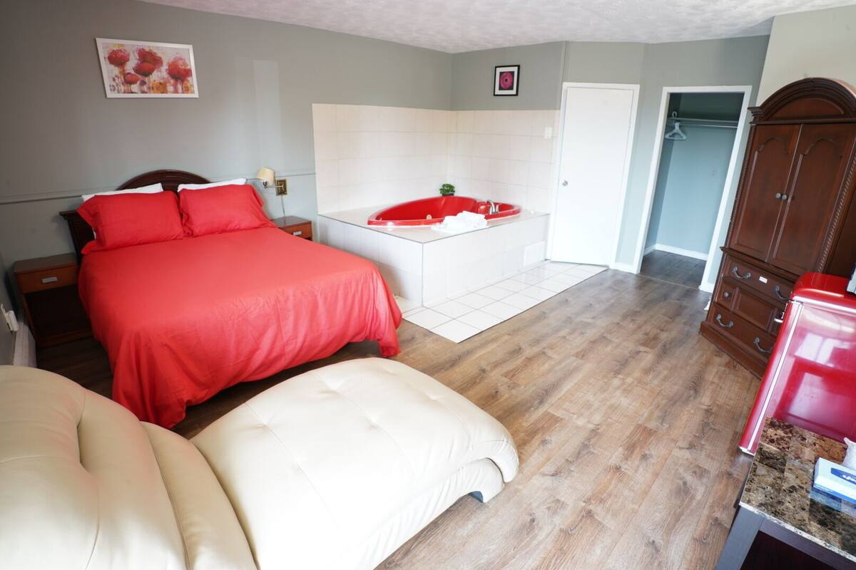 A bedroom with a large bed, a small bedside table, a red chair, a white bench, a bathroom with a red bathtub, and a wooden wardrobe.