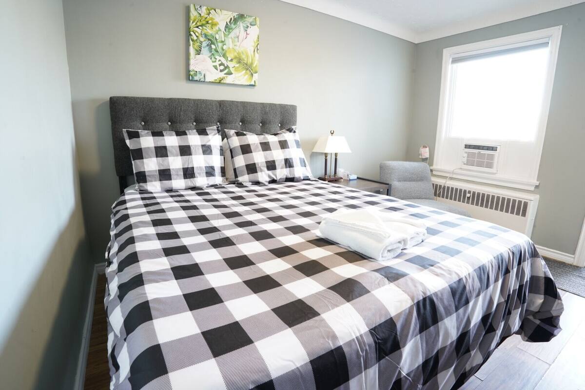 A neatly made bed with a checkered bedspread, a grey upholstered headboard, and a window with a view of the outdoors.