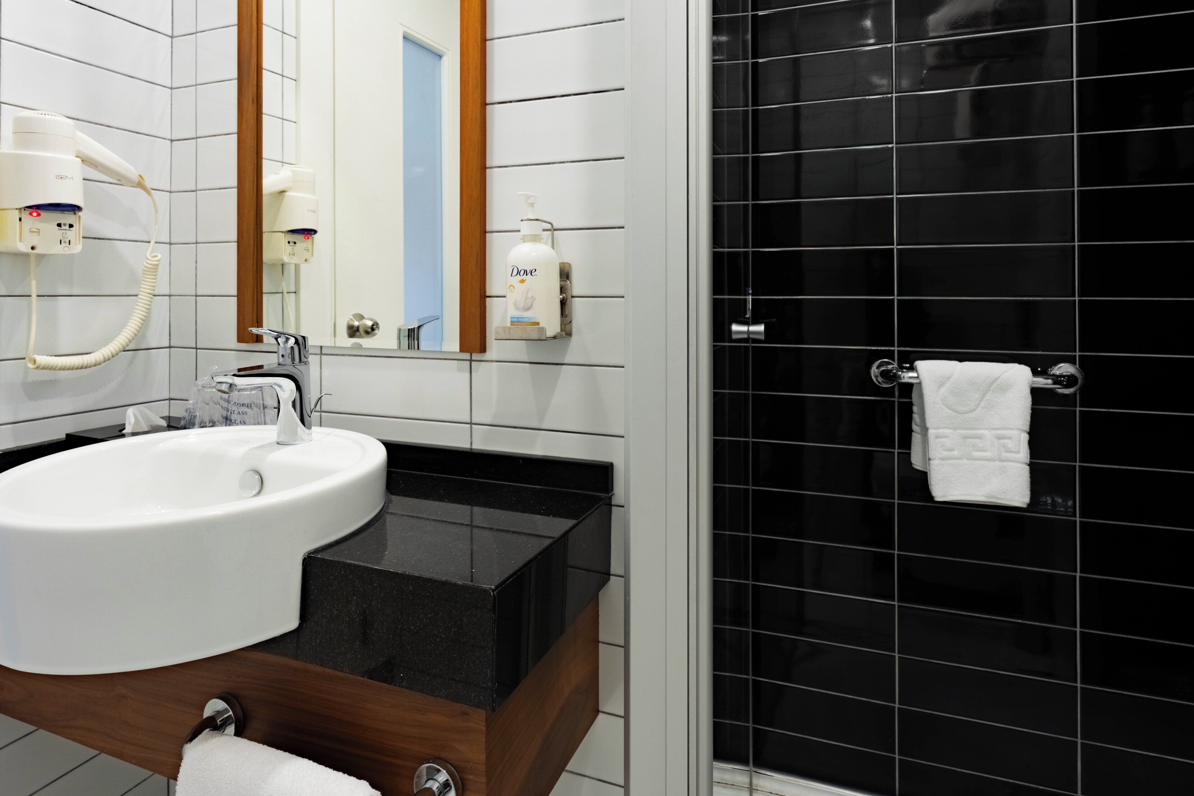 Un baño con un lavamanos blanco, un espejo, un secador de pelo de mano y una zona de ducha con azulejos negros.