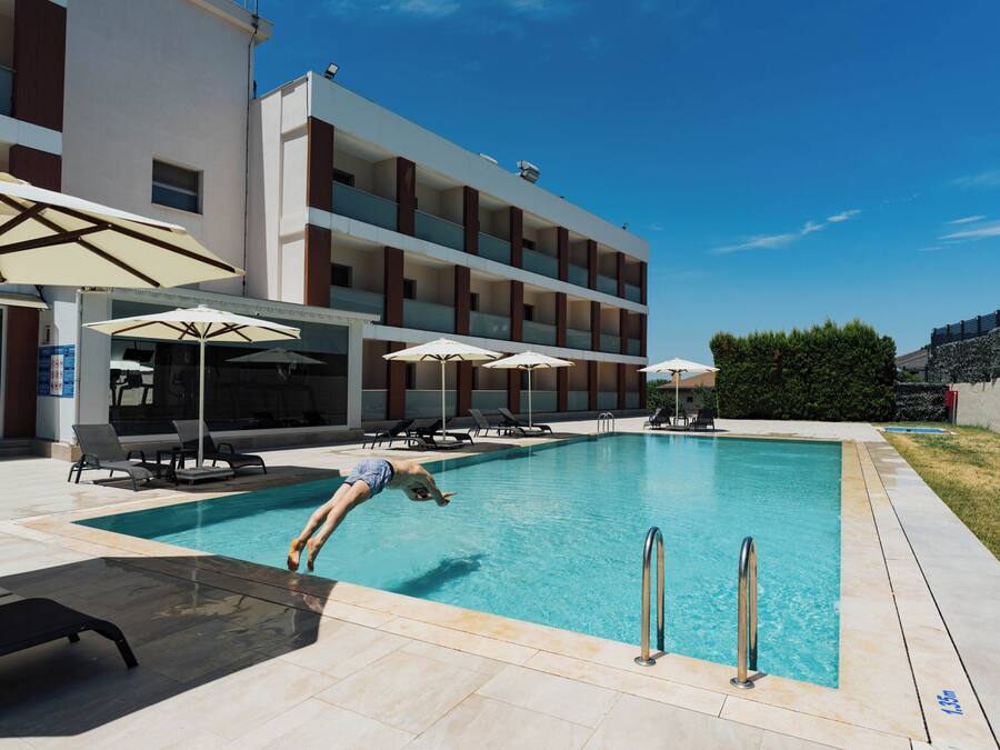 Una piscina con un trampolín, sillas de descanso, sombrillas y un edificio de varios pisos al fondo.