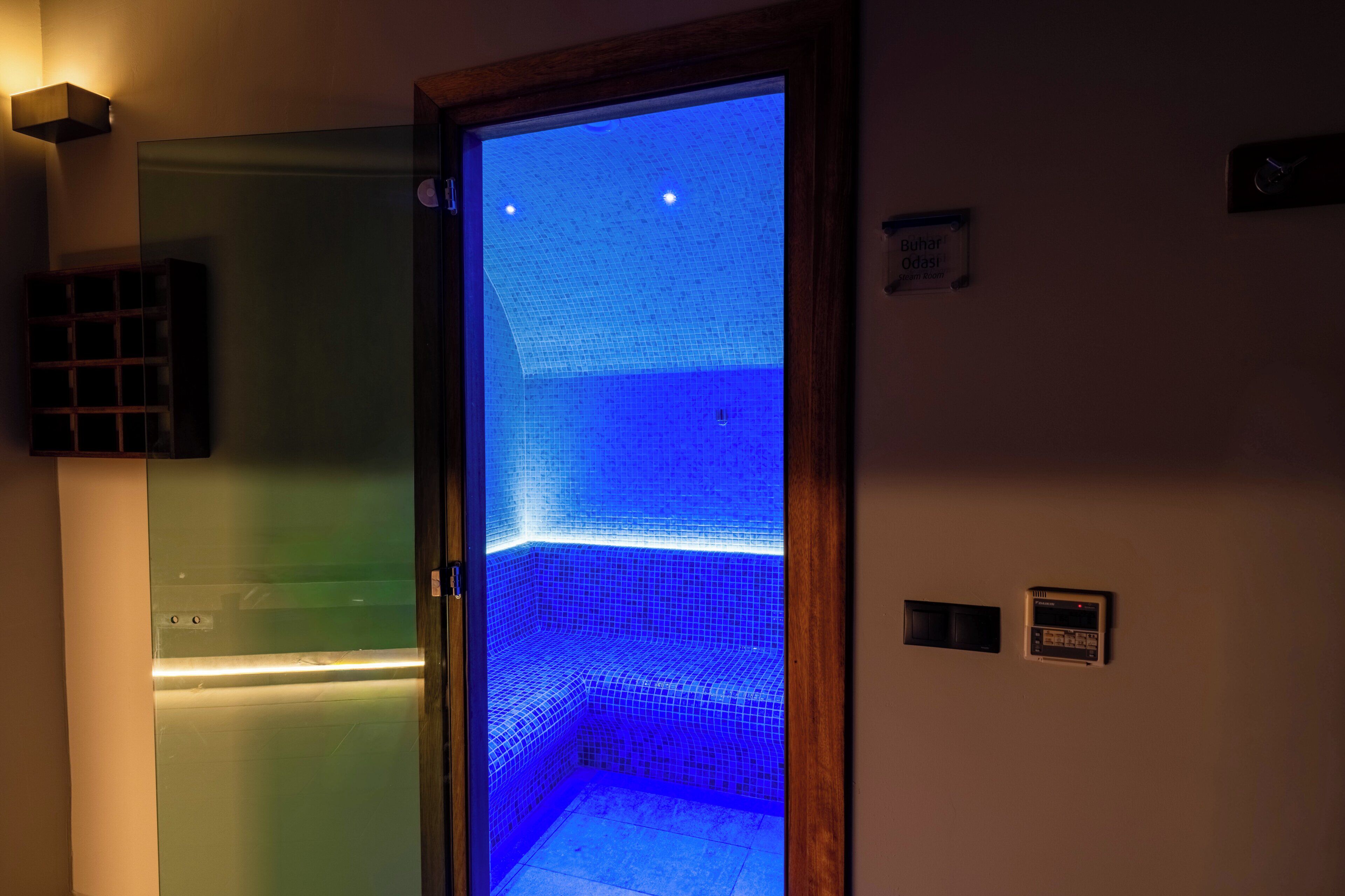 A room with a glass door leading to a blue-tiled area, a small sign, and a wall-mounted control panel.