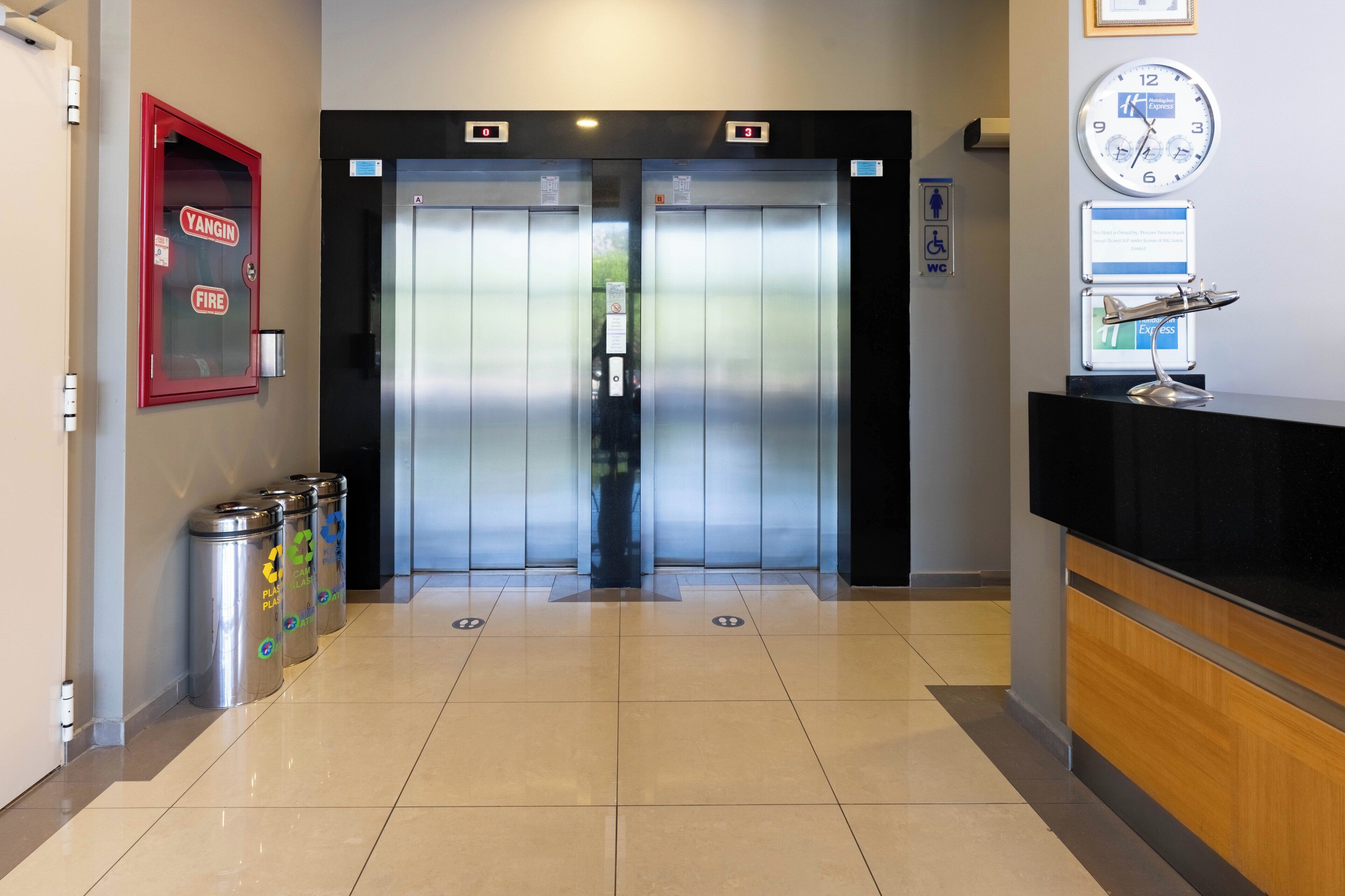 Un ascensor con pantallas digitales que indican los pisos A y B, un detector de incendios y un mostrador de recepción con un reloj.
