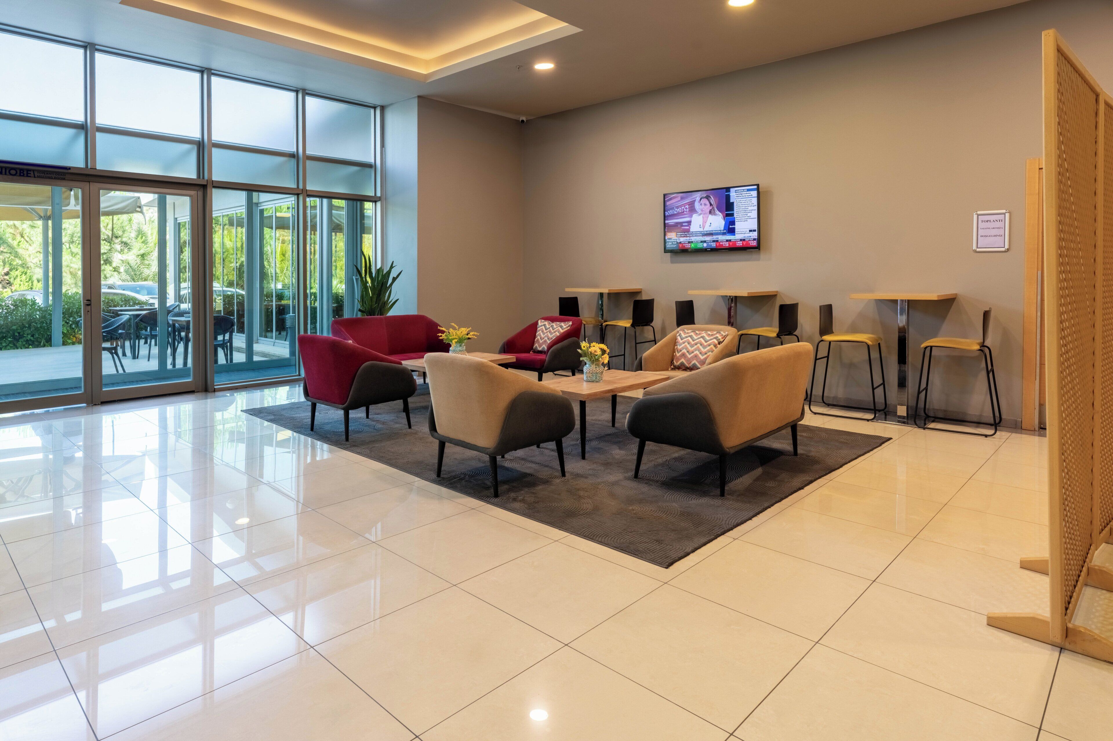A modern hotel lobby with a seating area, a flat-screen TV, and a large glass door leading to an outdoor area.