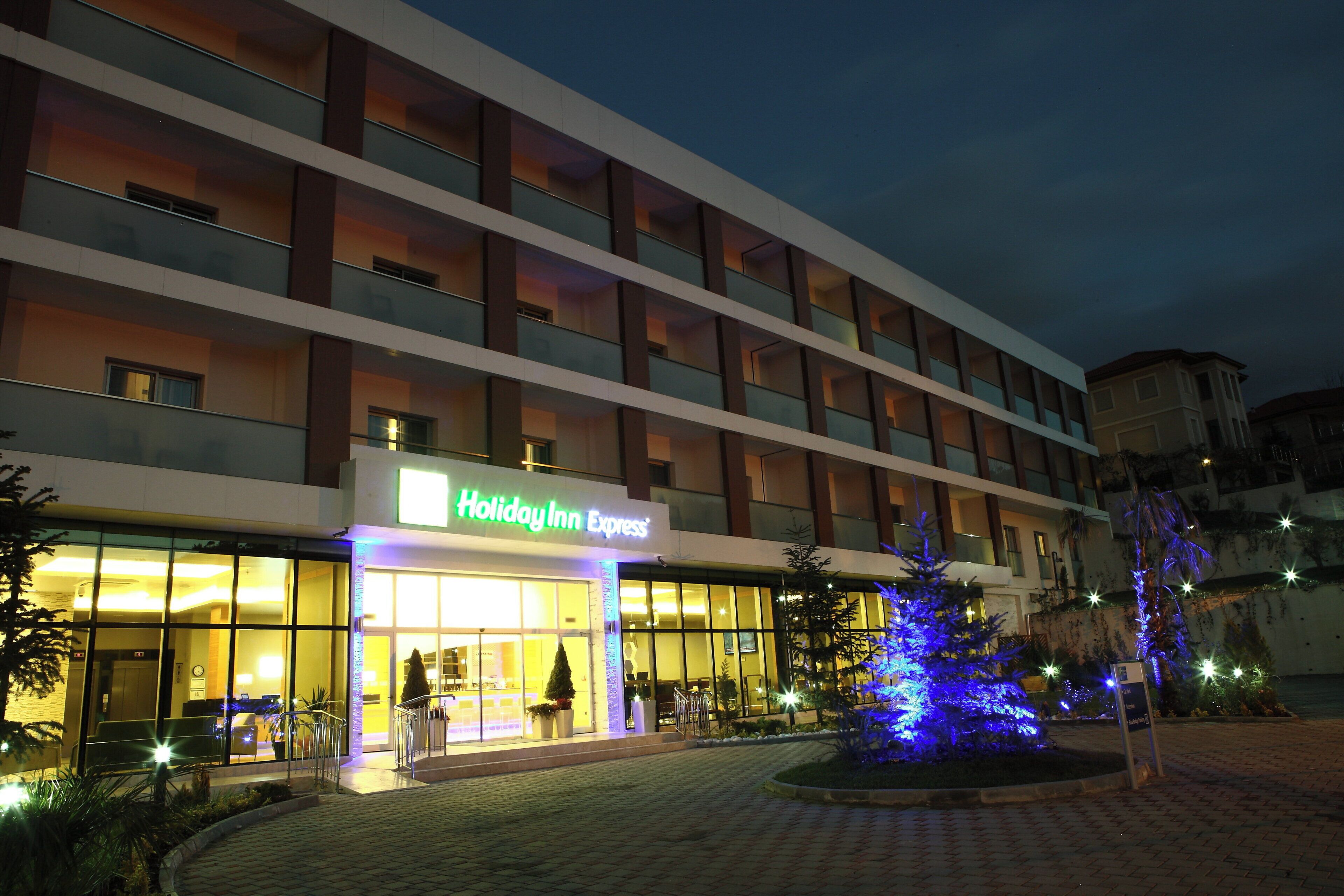 Edificio Holiday Inn Express con entrada bien iluminada, varios pisos y un área ajardinada con un árbol iluminado.