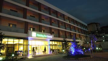 Edificio Holiday Inn Express con entrada bien iluminada, varios pisos y un área ajardinada con un árbol iluminado.