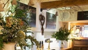 A bar area with wooden stools, a bar counter, and decorative plants. - The Bell at Hampton Poyle (Kidlington)