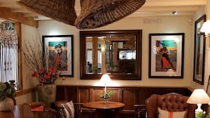 A cozy room with a wooden ceiling, a round table with a lamp, a cushioned bench, and framed pictures on the walls. - The Bell at Hampton Poyle (Kidlington)