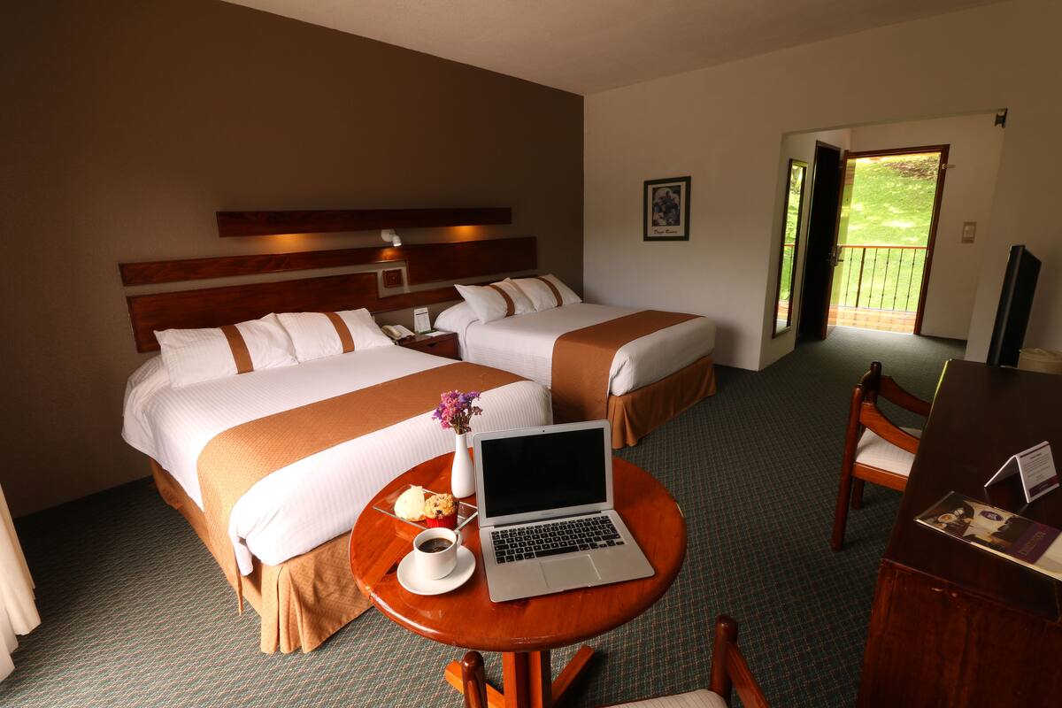 Habitación de hotel con dos camas, una laptop sobre una mesita, una silla y vista al exterior por una puerta.
