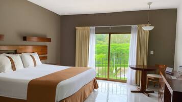 Habitación de hotel con una cama grande, un escritorio con silla, un balcón con cortinas y vistas a zonas verdes.