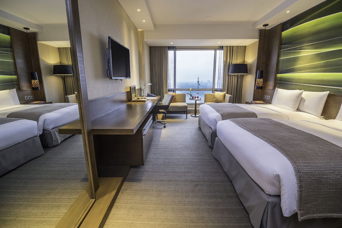 A hotel room with two beds, a desk, a chair, a TV, and a large window with a city view.