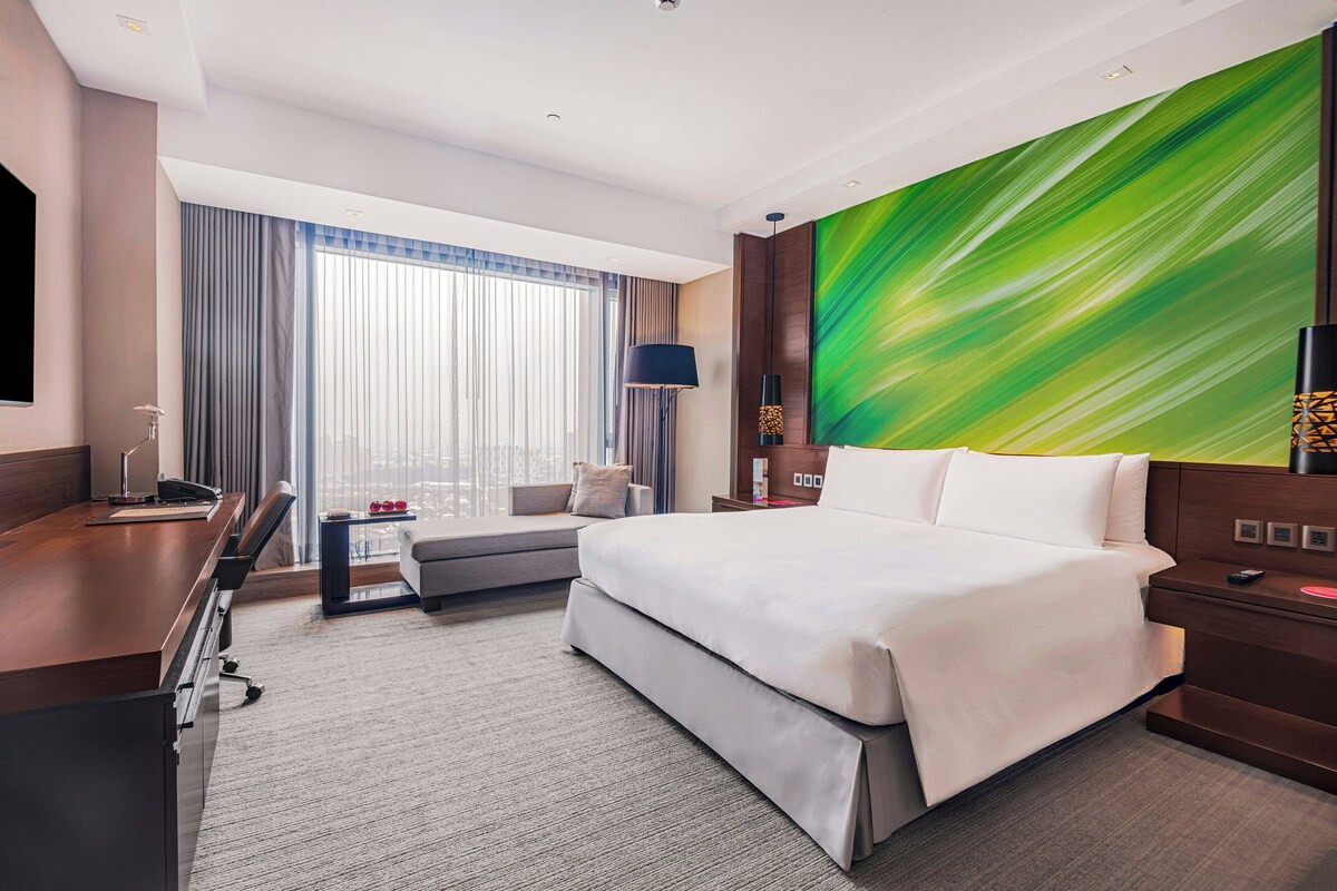 Habitación de hotel con cama, escritorio, silla, sofá, ventana grande con cortinas, televisor montado en la pared y un cuadro.