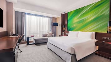 A hotel room with a bed, a desk, a chair, a sofa, a large window with curtains, a wall-mounted TV, and a painting.