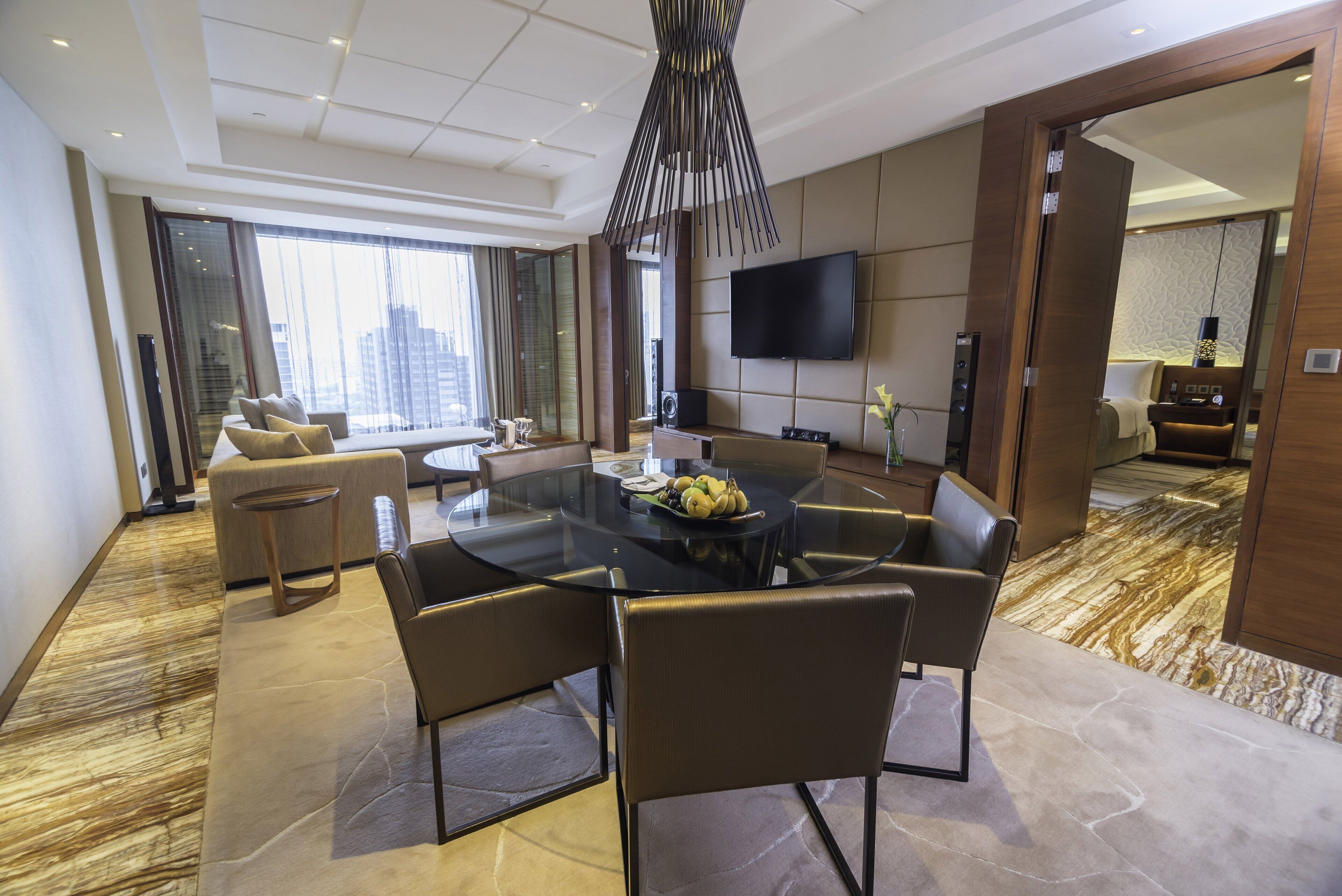 A modern hotel room with a dining area, a sofa, a TV, and a view of the city.