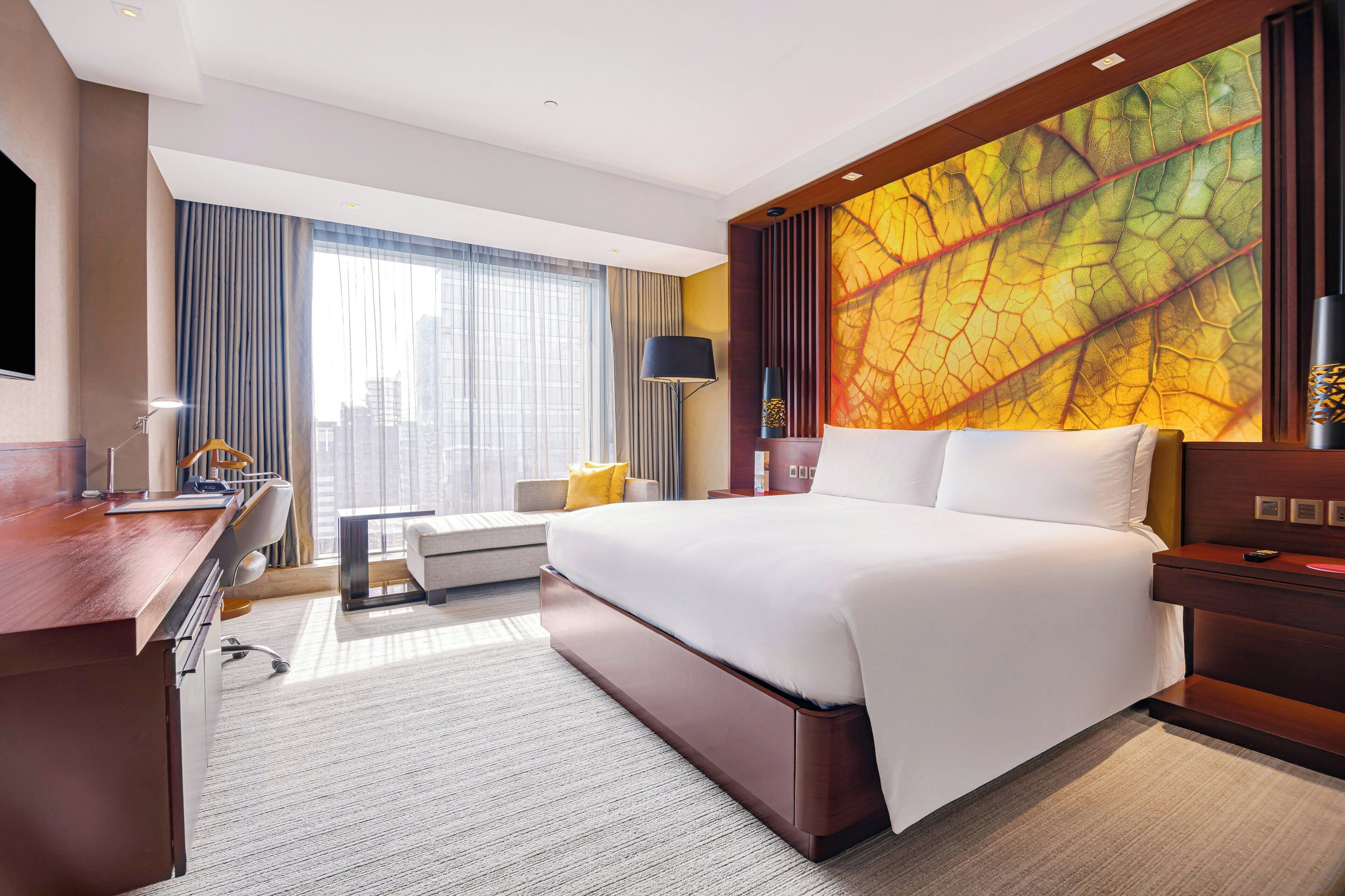 A hotel room with a large bed, a desk, a chair, a sofa, a large window with curtains, and a wall with a leaf pattern.