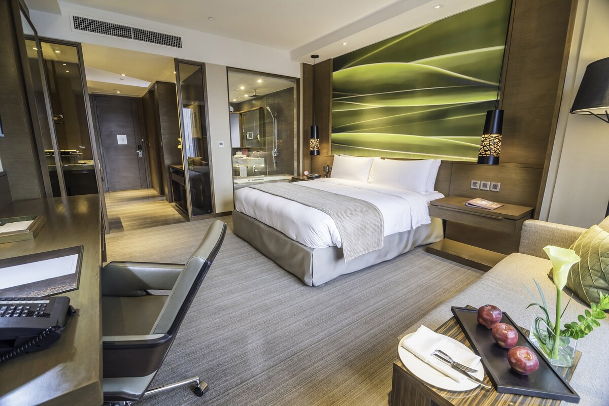 Una habitación de hotel moderna con una cama grande, un escritorio con teléfono, una zona de estar con mesa y sillas, y un baño visible a través de una puerta de vidrio.
