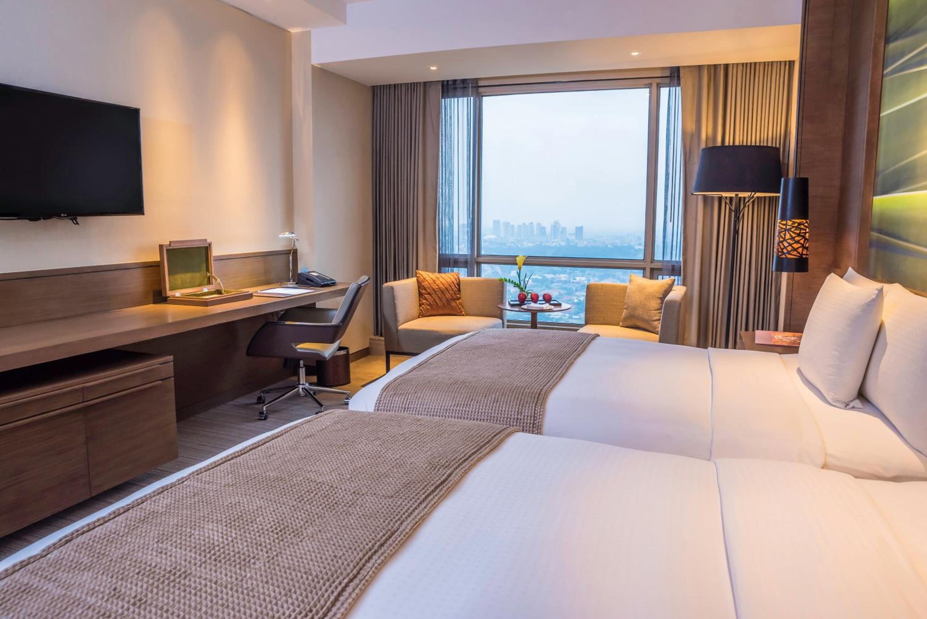 A hotel room with two beds, a desk, a TV, and a view of the city.