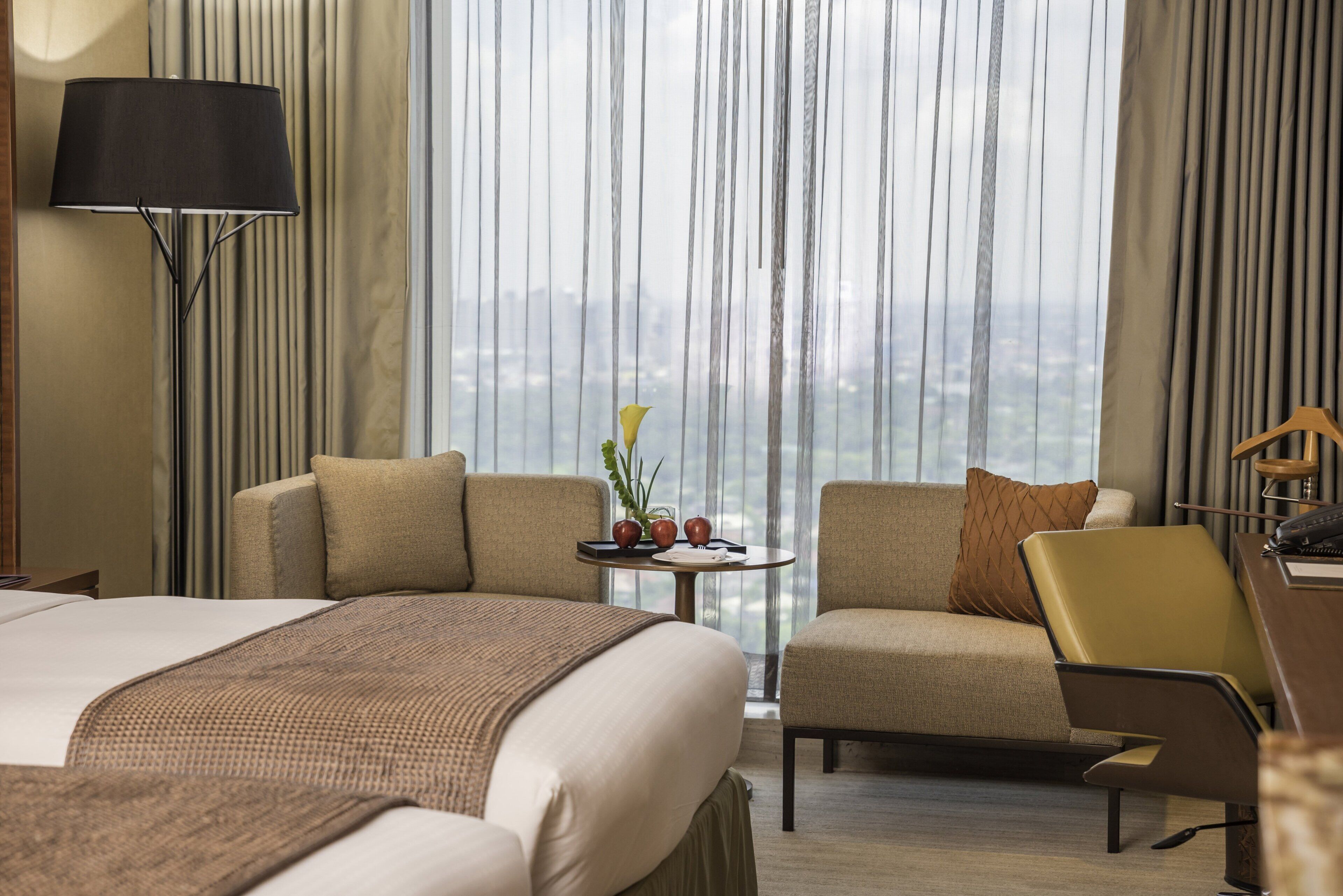 A hotel room with a bed, two armchairs, a small table with a plant, and a view of a cityscape through large windows.