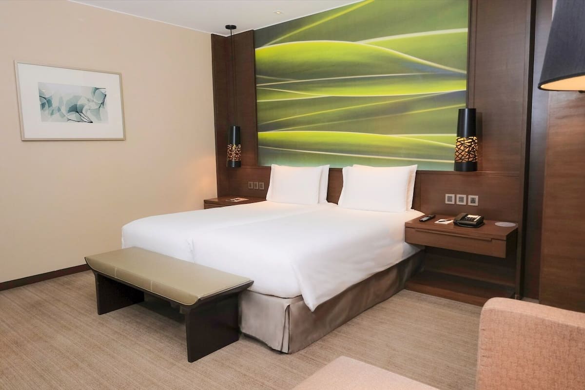 A hotel room with a large bed, a bench, a desk, and a wall with a green abstract painting.