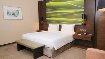A hotel room with a large bed, a bench, a desk, and a wall with a green abstract painting.
