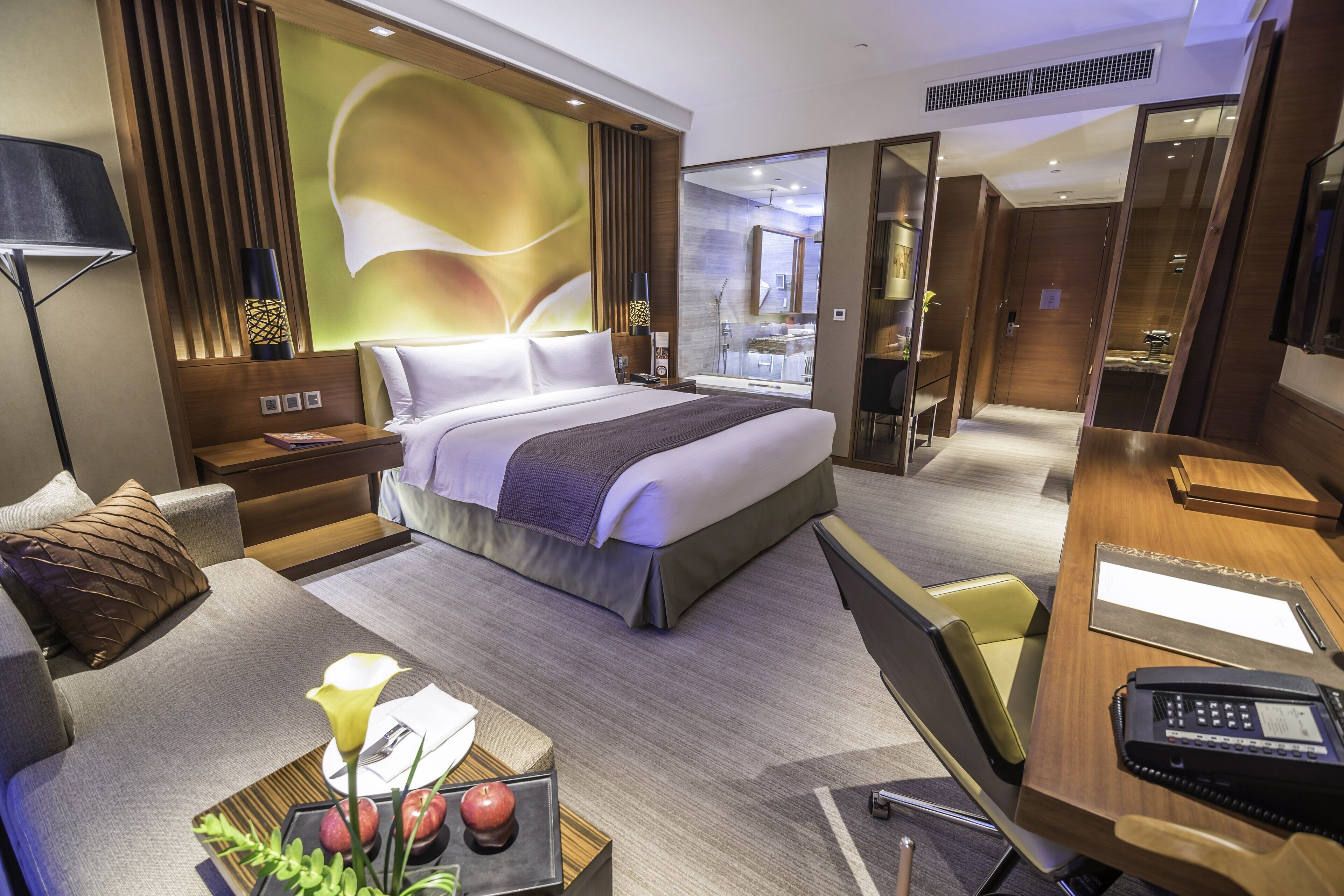 A modern hotel room with a large bed, a desk with a telephone, a seating area with a sofa and a coffee table with fruits and a flower.