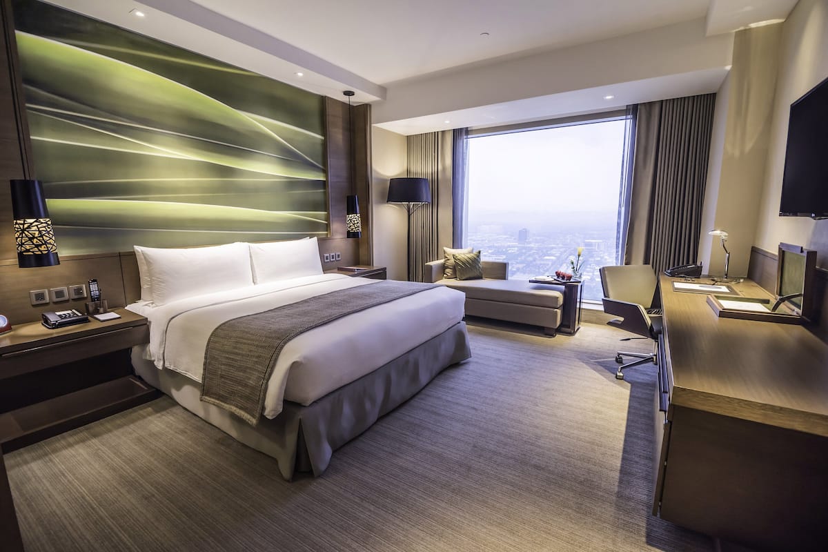 A modern hotel room with a large bed, a desk, a chair, a sofa, and a large window offering a city view.
