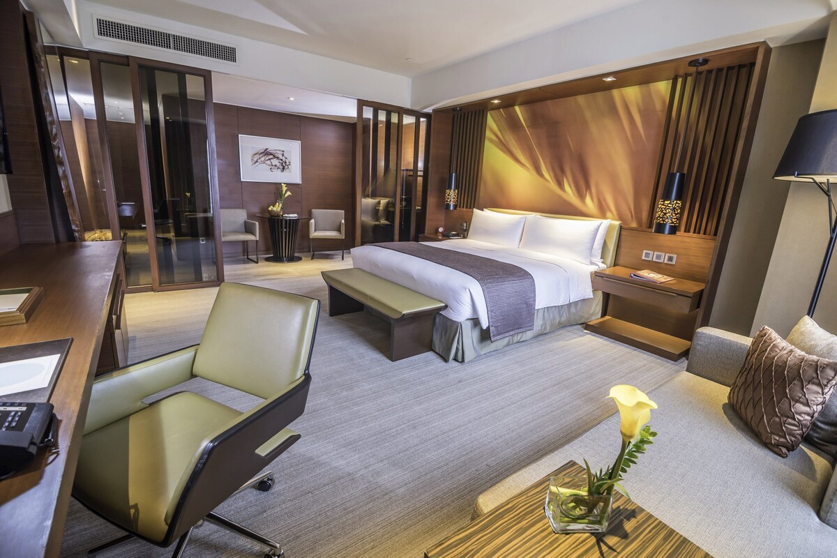 Habitación de hotel con cama, escritorio, sillón, sofá y mesita de noche con un florero.