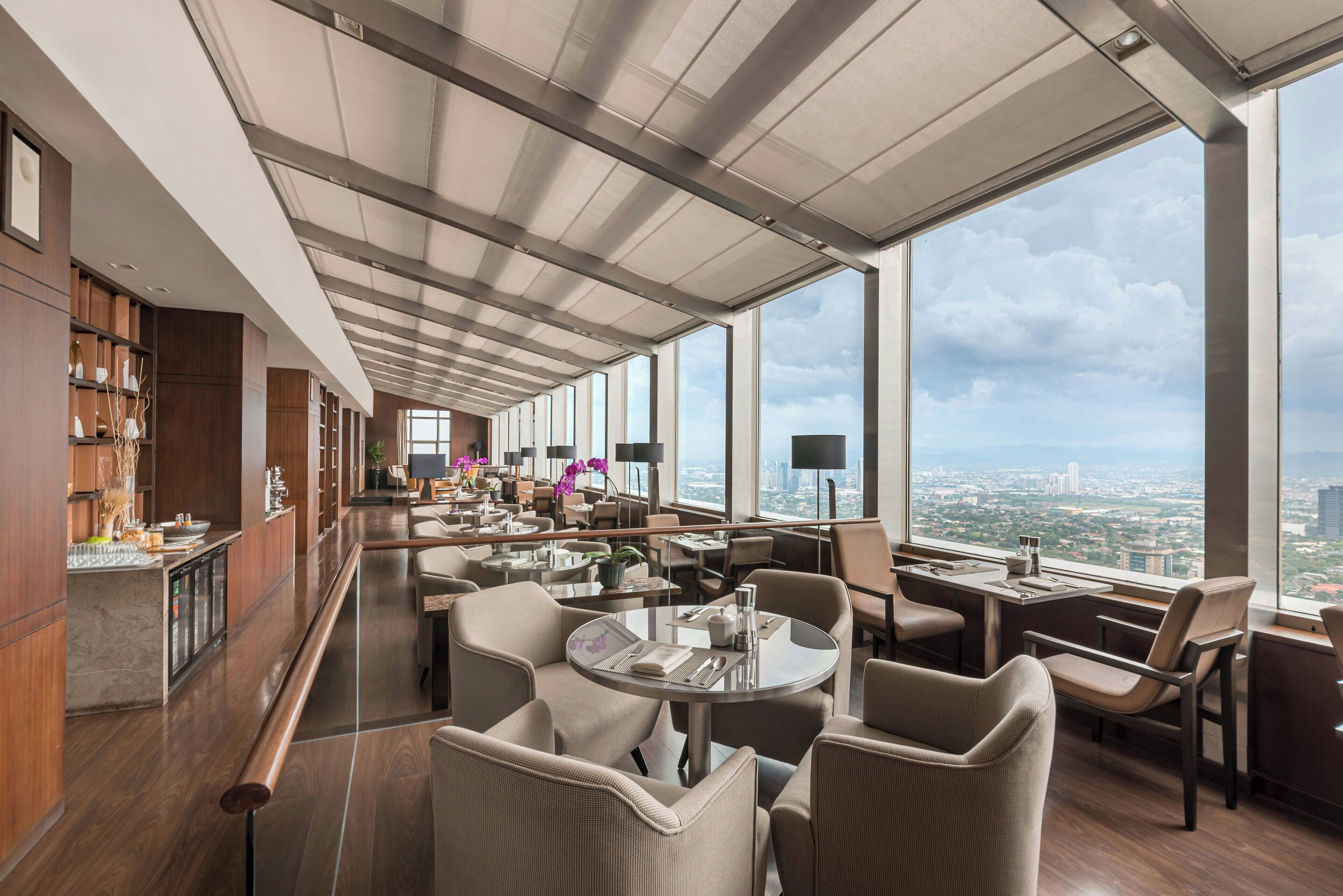 A modern hotel lobby with a high ceiling, large windows offering city views, and a bar area with wooden cabinets.