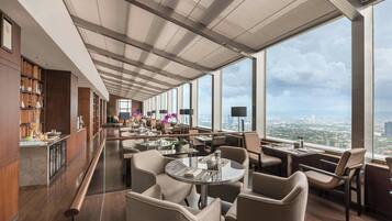 A modern hotel lobby with a high ceiling, large windows offering city views, and a bar area with wooden cabinets.