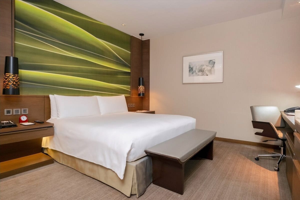 A hotel room with a large bed, a desk with a chair, a bedside table with a lamp, and a large abstract painting on the wall.