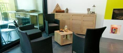 A modern living room with wicker furniture, a small wooden table with snacks, and a kitchen area visible through a glass door.