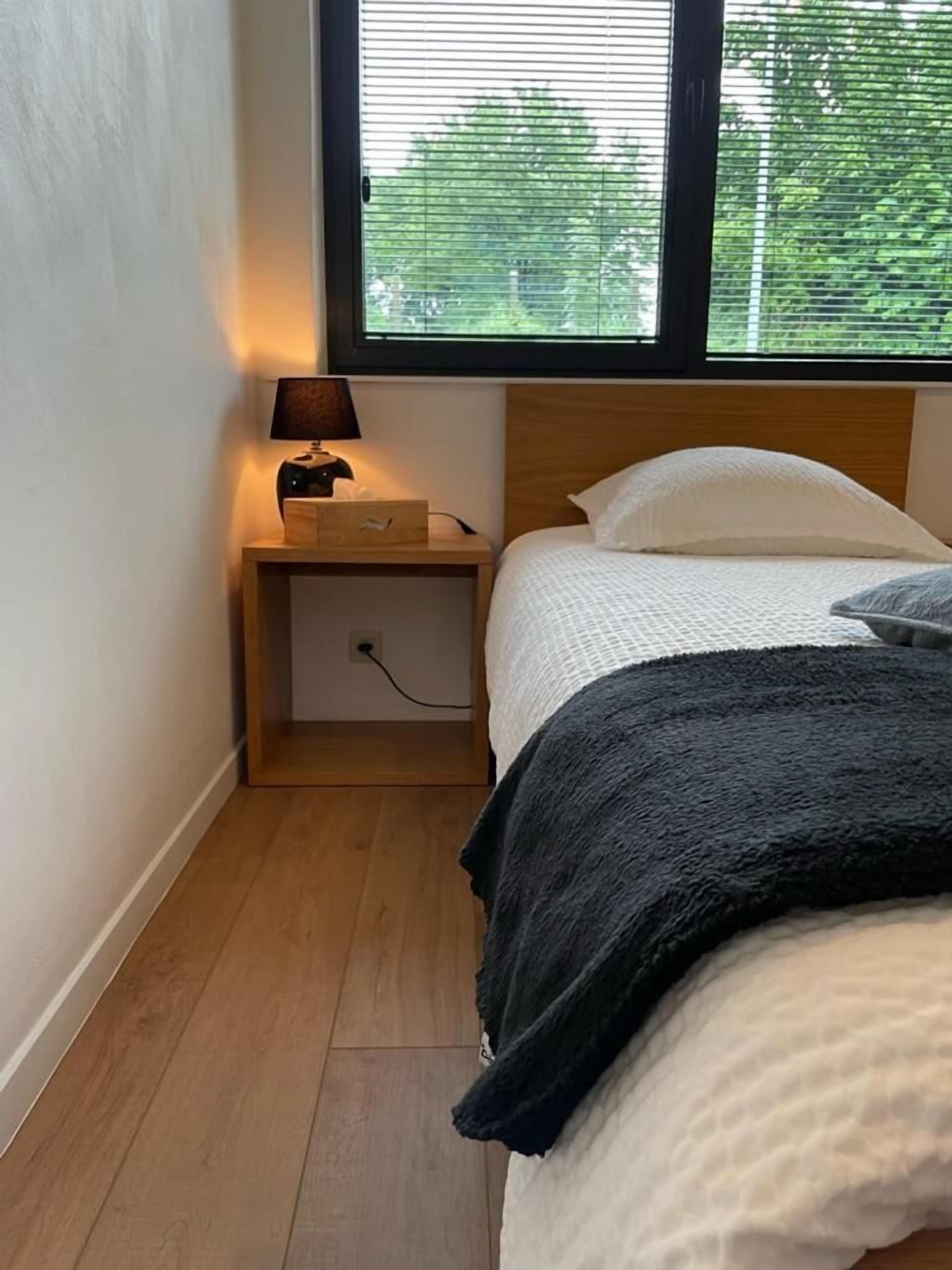 Un dormitorio con una cama, una mesita de noche con lámpara y un ventanal con persianas.