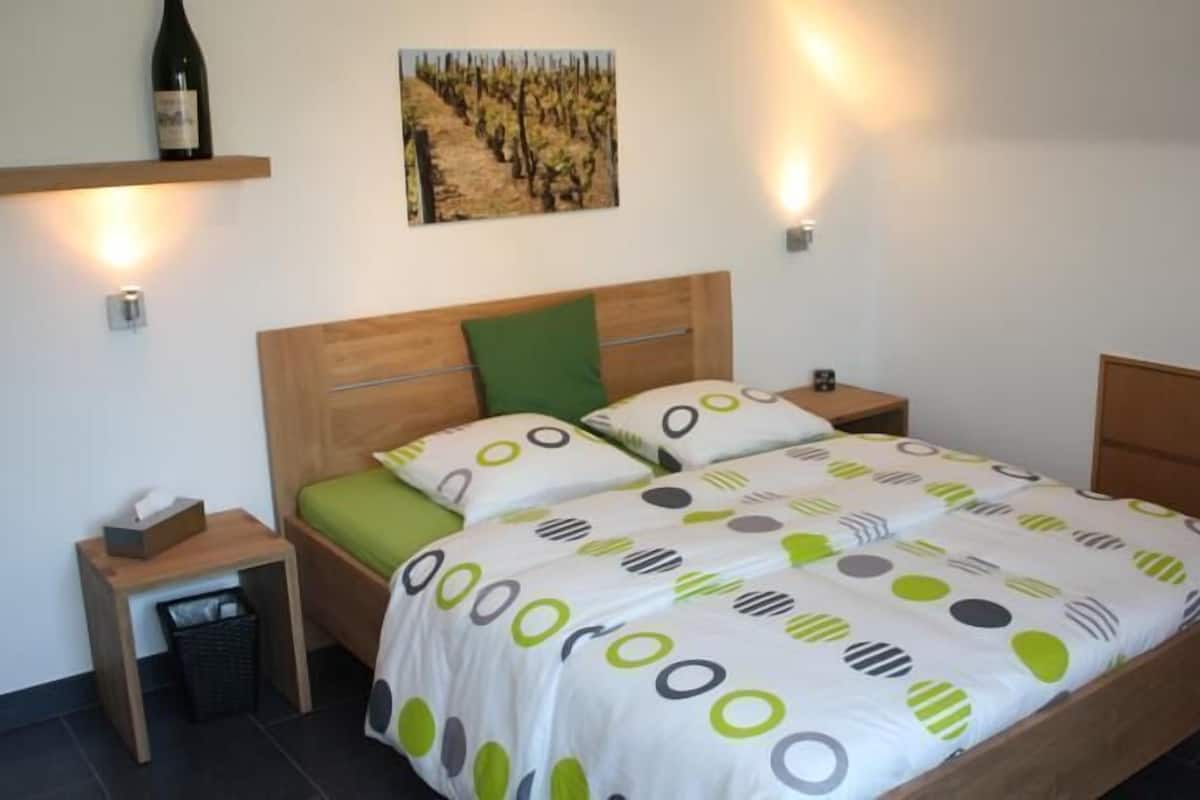 Ein Schlafzimmer mit einem hölzernen Bett, grün-weißer Bettwäsche, einem Nachttisch mit Lampe, einem Mülleimer und einem an der Wand befestigten Regal mit einer Flasche.