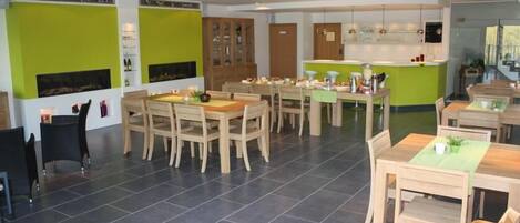 A modern dining area with wooden tables and chairs, a kitchen in the background, and green accent walls.