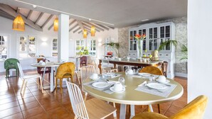 Un comedor amplio con una gran mesa redonda dispuesta para cuatro, rodeada de sillas. Hay lámparas colgantes sobre la mesa, y se ve una zona de cocina al fondo.