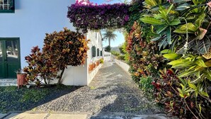 Un camino adoquinado que lleva a un edificio blanco con contraventanas verdes y un balcón cubierto de flores moradas.