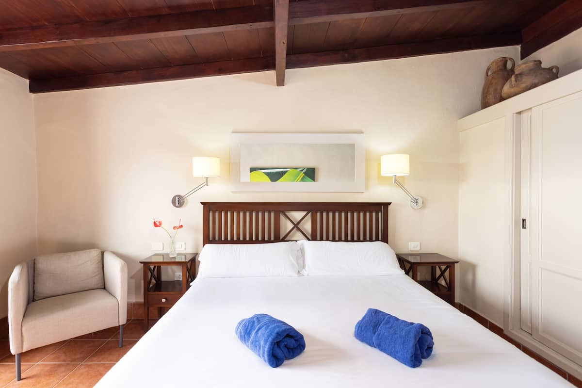 A hotel room with a bed, a chair, a nightstand, a lamp, and a painting on the wall.