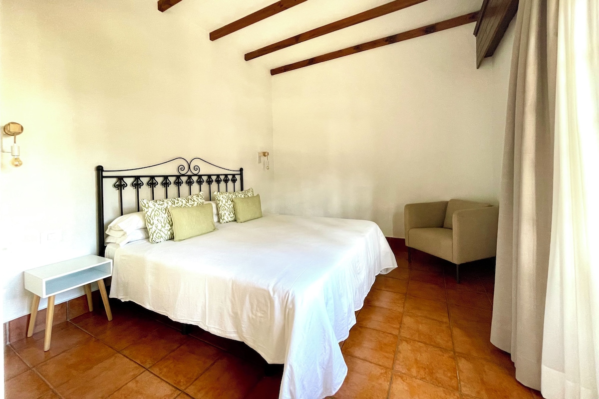 Un dormitorio con cama, mesita de noche, una silla y ventana con cortinas.
