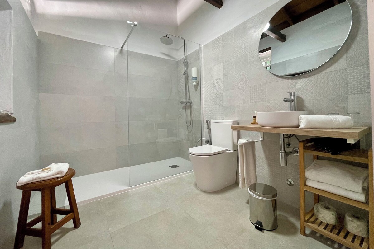 Un baño moderno con ducha de vidrio, inodoro blanco, lavamanos con espejo y un taburete de madera con una toalla.