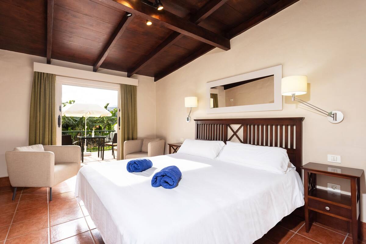 A hotel room with a large bed, two bedside lamps, a wooden headboard, a small bedside table, a chair, and a view of greenery outside.