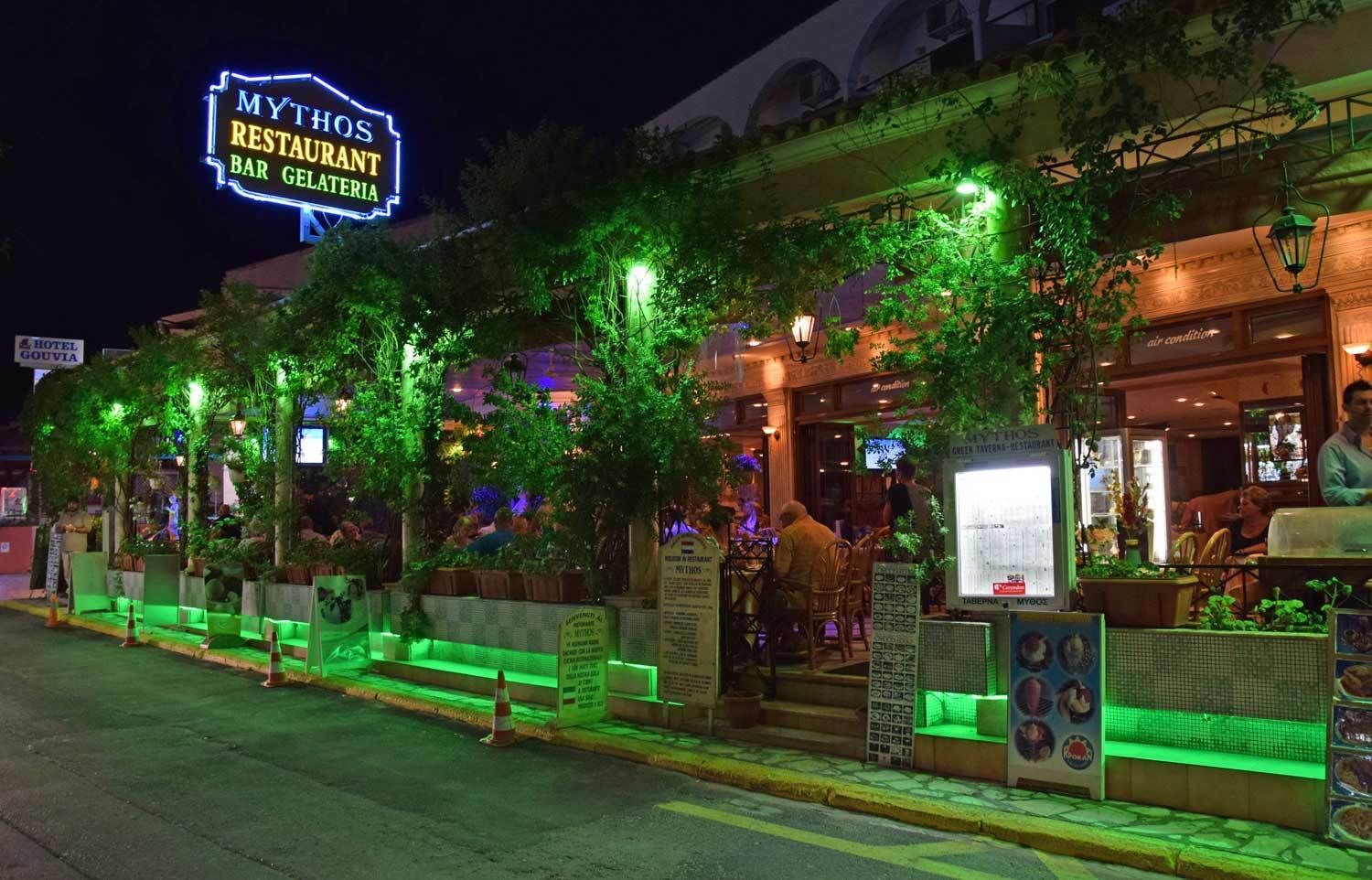 A restaurant with outdoor seating, green plants, and a neon sign.