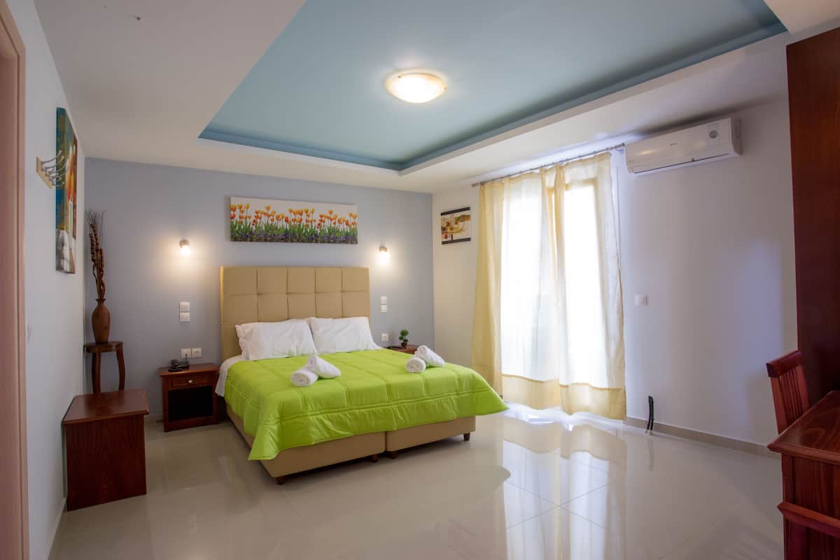 A hotel room with a bed, a nightstand, a small table, a chair, a painting, and an air conditioning unit.